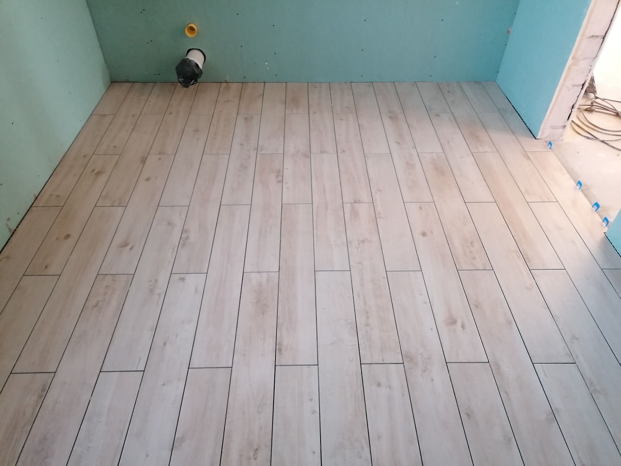 Ułożona podłoga z płytek imitujących drewno w odcieniach beżu i bieli, widoczne niebieskie kliny dystansowe, ściany w surowym stanie z instalacją kanalizacyjną.