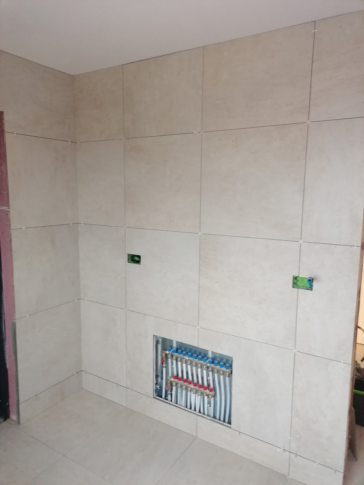 Ściana i podłoga wyłożone beżowymi, kwadratowymi płytkami, widoczne instalacje hydrauliczne w otworze ściennym, gniazdka elektryczne w ścianie, etap prac wykończeniowych.