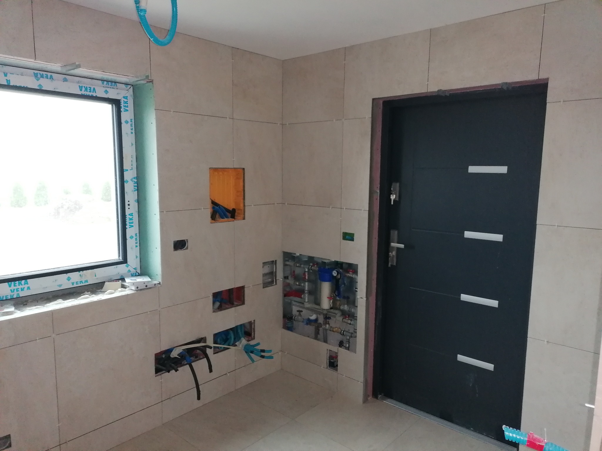 Łazienka w trakcie wykańczania: jasne płytki na ścianach i podłodze, okno z profilem VEKA, widoczne instalacje hydrauliczne w otworach w ścianie, czarne drzwi z poziomymi wstawkami, niebieskie...