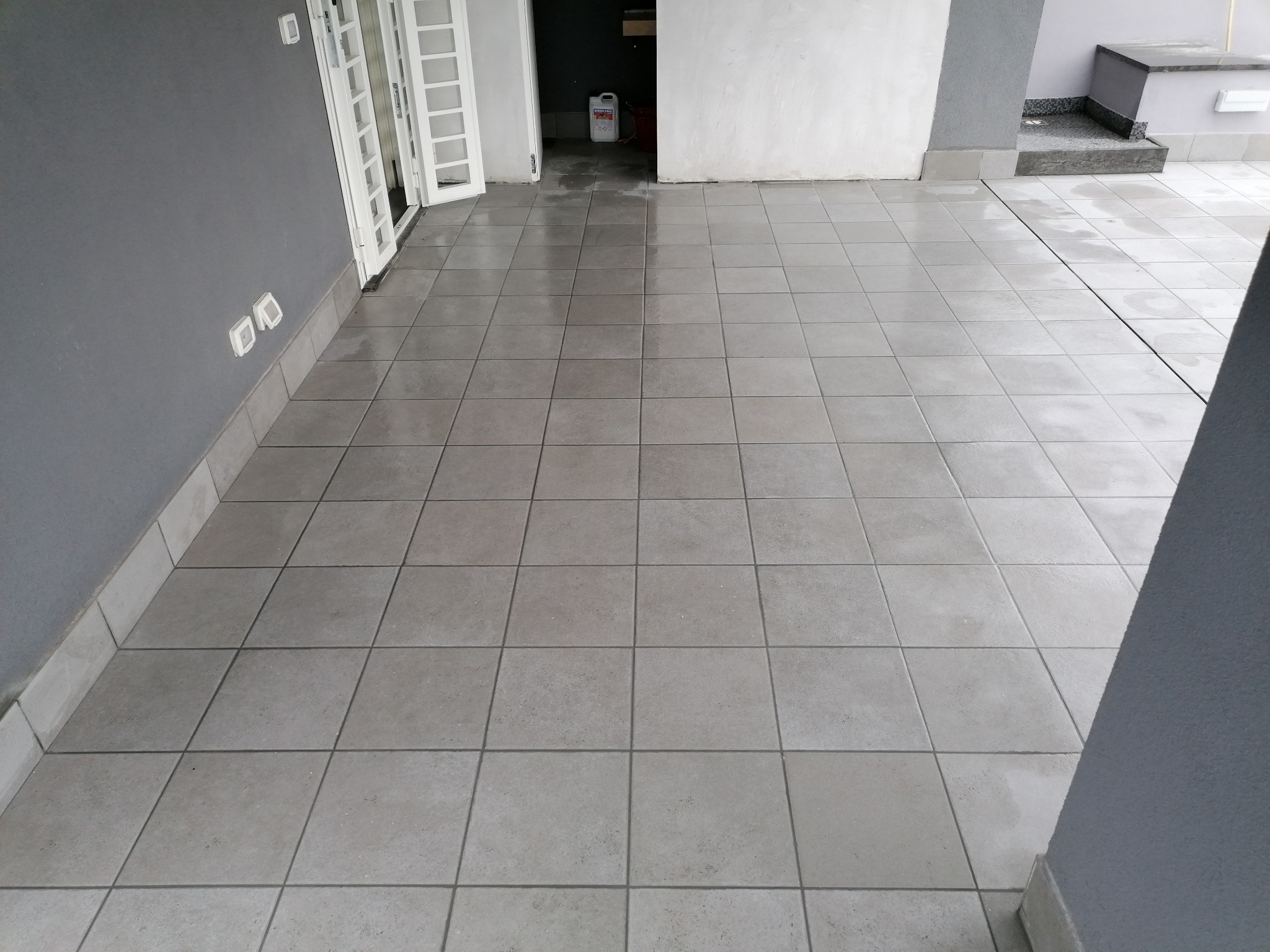 Szare, kwadratowe płytki podłogowe po remoncie, widoczne ślady wilgoci, białe drzwi balkonowe w tle, preparat do czyszczenia w rogu.