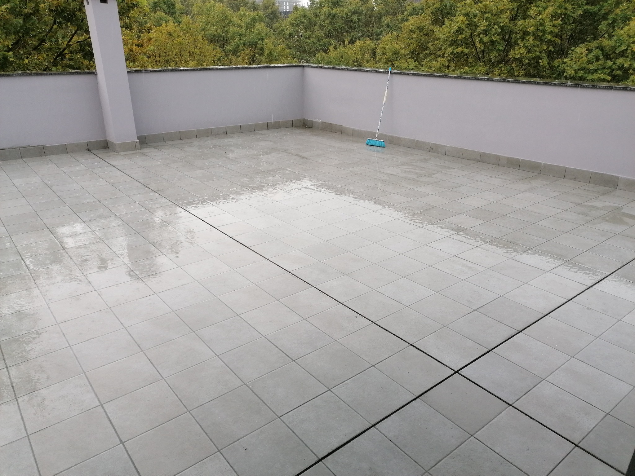 Szary taras z nowymi płytkami, mokry po umyciu, z widoczną szczotką do zamiatania przy ścianie i drzewami w tle.