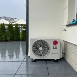 Zewnętrzna jednostka klimatyzacji LG Dual Inverter zamontowana na szarym, mokrym tarasie, z widocznym kranem na ścianie i tujami w tle.
