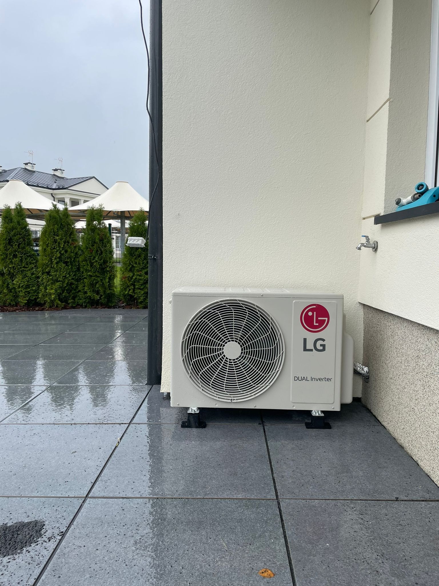 Zewnętrzna jednostka klimatyzacji LG Dual Inverter zamontowana na szarym, mokrym tarasie, z widocznym kranem na ścianie i tujami w tle.