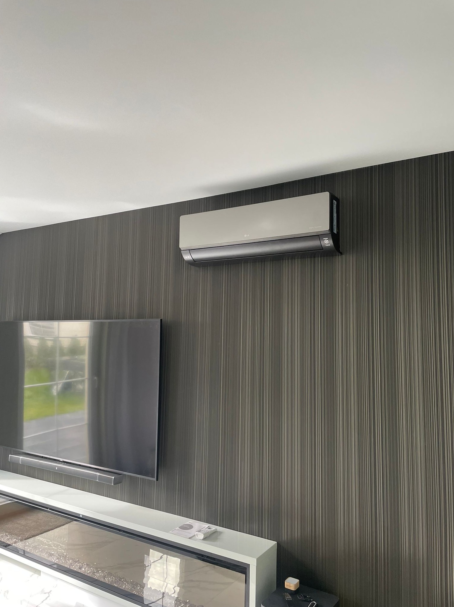 Nowoczesny klimatyzator ścienny LG Dual Inverter zamontowany nad telewizorem w salonie z ciemną tapetą w pionowe paski.
