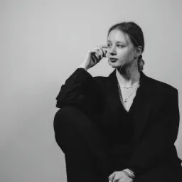 Czarno-białe zdjęcie kobiety w czarnym garniturze, siedzącej ze skrzyżowanymi nogami i zamyślonym spojrzeniem, z biżuterią: perłowy naszyjnik i bransoletka, kolczyki koła.