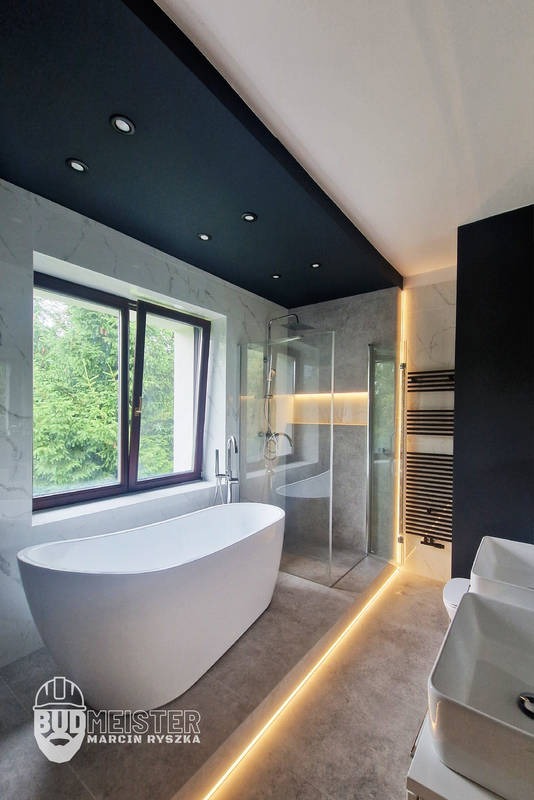 Nowoczesna łazienka z wolnostojącą wanną przy oknie, szklaną kabiną prysznicową i podświetleniem LED. Marmurowe płytki i ciemny sufit z oświetleniem punktowym.
