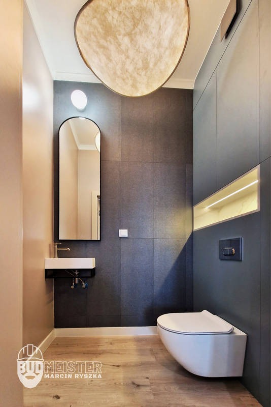 Nowoczesna łazienka z podwieszaną toaletą, umywalką i dużym lustrem w czarnej ramie. Ciemne płytki na ścianach, drewniana podłoga i designerska lampa sufitowa.