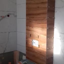 Montaż płytek imitujących drewno na obudowie stelaża podtynkowego w łazience, widoczne krzyżyki dystansowe i otwór na przycisk spłukujący.