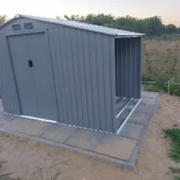 Szary metalowy domek narzędziowy z otwartym wejściem, ustawiony na betonowej podstawie z kostki, widok na krajobraz z zielenią i ogrodzeniem w tle.