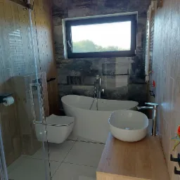 Nowoczesna łazienka z wolnostojącą wanną, umywalką nablatową i kabiną prysznicową ze szklanymi drzwiami, wykończona płytkami imitującymi drewno i kamień, z oknem w czarnej ramie.