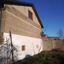 Dom z częściowo ocieploną elewacją: dolna część ściany pokryta białym styropianem, górna część z szarego pustaka, brązowe okna, w tle widoczny płot z betonowych płyt.