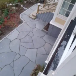 Nieregularna, szara kamienna ścieżka prowadząca do schodów z drewnianą balustradą, widok z góry z fragmentem rynny w kadrze.