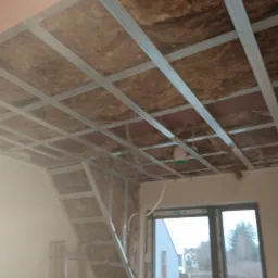 Poddasze z widoczną konstrukcją metalową sufitu podwieszanego i izolacją z wełny mineralnej, okno w tle.