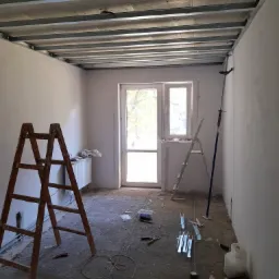 Wnętrze w trakcie montażu metalowej konstrukcji sufitu podwieszanego z płyt kartonowo-gipsowych, widoczne drabiny, narzędzia i materiały budowlane na podłodze.