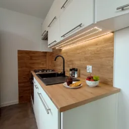 Nowoczesna, jasna kuchnia z drewnianym blatem i panelami ściennymi, oświetleniem LED pod szafkami, czarnym zlewem i kuchenką gazową. Na blacie misa z owocami i talerz z ciastem.