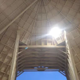 Wnętrze drewnianej konstrukcji dachu z widocznym niebem przez otwór, oświetlone mocnym światłem w centrum kadru.