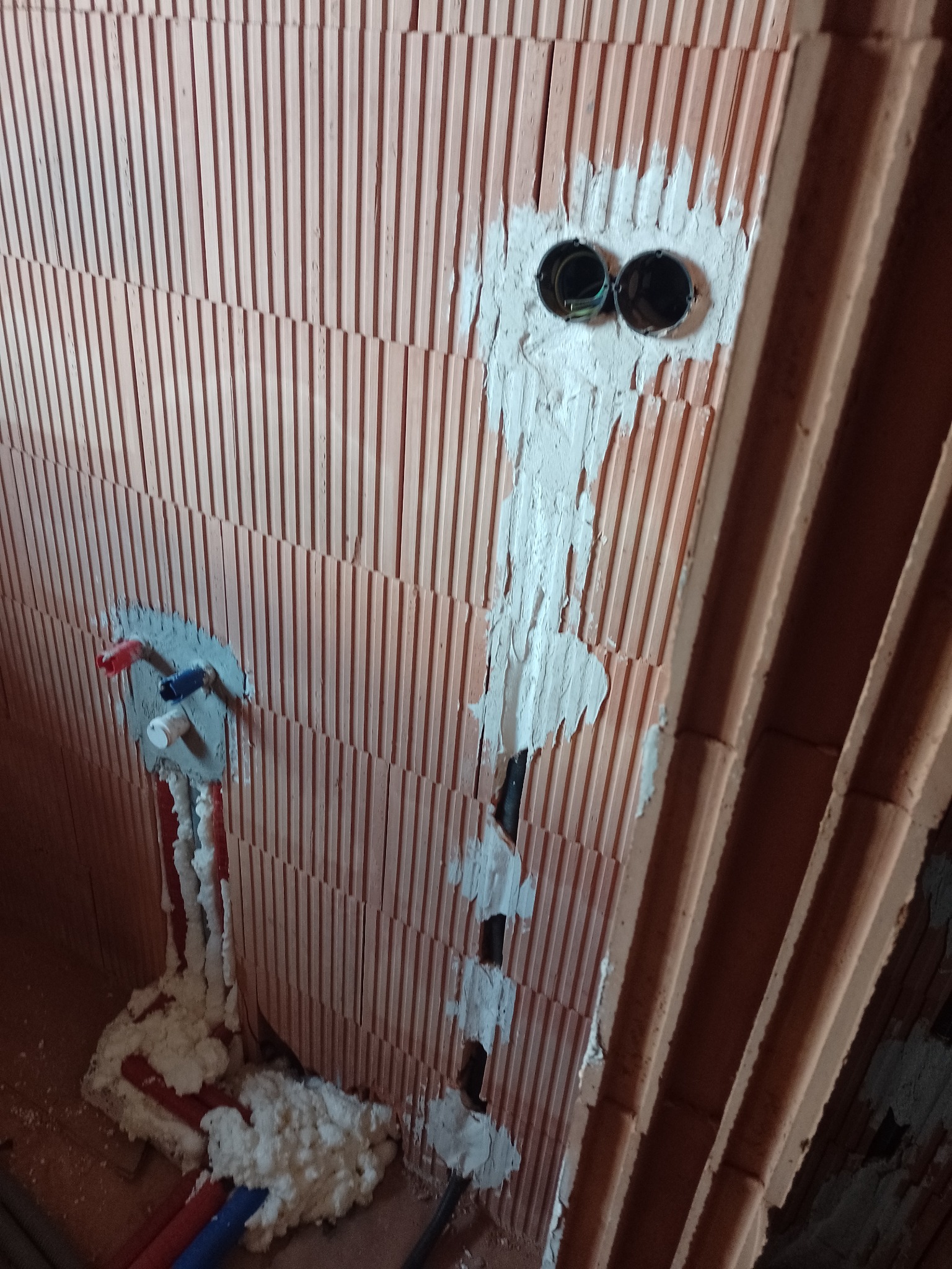 Fragment ściany z pustaków ceramicznych z widocznymi puszkami instalacyjnymi zalane gipsem oraz instalacją wodną zabezpieczoną pianką montażową.