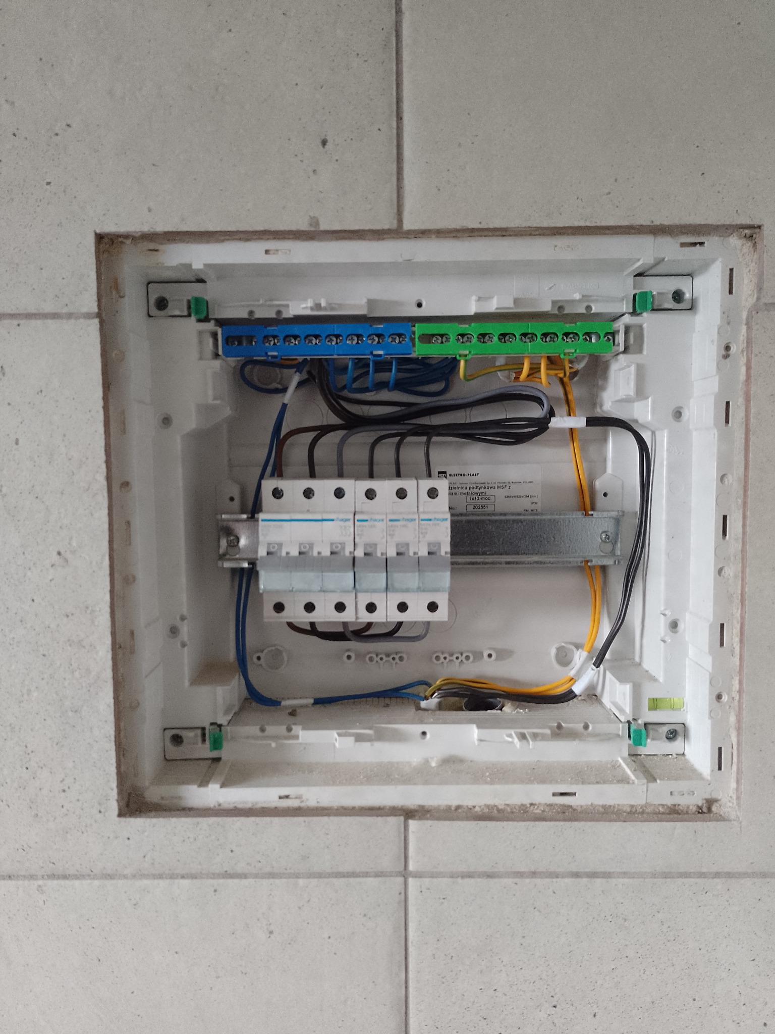 Otwarta skrzynka elektryczna podtynkowa z bezpiecznikami, listwami zaciskowymi i okablowaniem, zamontowana w ścianie wyłożonej płytkami ceramicznymi, widoczna poziomica.