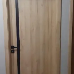 Zamknięte, drewniane drzwi wewnętrzne z czarnym, pionowym paskiem i czarną klamką na tle szarej ściany i fragmentu drugich drzwi.