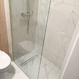 Nowoczesna kabina prysznicowa z odpływem liniowym, wykończona białymi płytkami imitującymi marmur, widoczny fragment toalety.