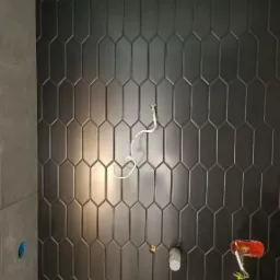 Ściana wyłożona czarnymi, podłużnymi płytkami o heksagonalnym kształcie, z widocznymi instalacjami elektrycznymi i wodnymi w trakcie montażu.