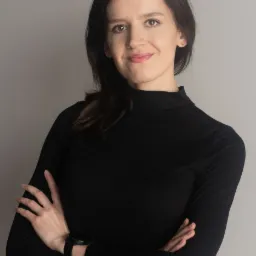 Portret uśmiechniętej kobiety w czarnym golfie, z rękami skrzyżowanymi na piersiach, na jednolitym szarym tle.