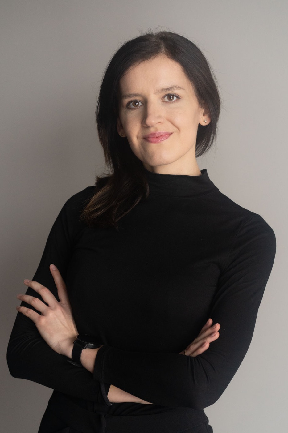 Portret uśmiechniętej kobiety w czarnym golfie, z rękami skrzyżowanymi na piersiach, na jednolitym szarym tle.