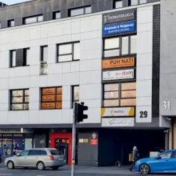 Ulica Warszawska w Kielcach z widocznymi budynkami, szyldami firm i zaparkowanymi samochodami. Na jednym z budynków widnieje numer 29 oraz szyldy różnych firm, w tym prawdopodobnie kancelarii...