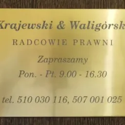Złota tabliczka informacyjna kancelarii radców prawnych Krajewski & Waligórski w Kielcach, z godzinami otwarcia i numerami telefonów, umieszczona na ciemnym, drewnianym tle.