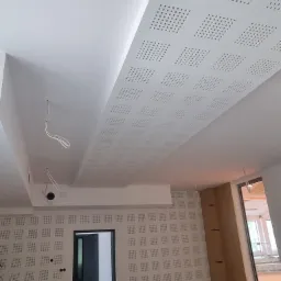 Pokój z wykończonym sufitem i ścianami z perforowanych paneli gipsowych w kwadratowy wzór, widoczne kable wystające z sufitu i otwarte drzwi do kolejnego pomieszczenia.