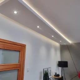 Nowoczesne wnętrze z podwieszanym sufitem z oświetleniem LED i punktowymi lampami, widoczny fragment drewnianych drzwi i minimalistyczna komoda z dekoracjami.