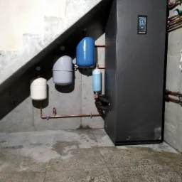 Czarna pompa ciepła z zbiornikami wyrównawczymi pod betonowymi schodami w piwnicy, widoczne miedziane rury instalacji.