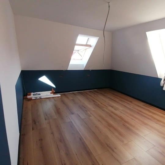 Poddasze z drewnianą podłogą w trakcie prac wykończeniowych, ściany pomalowane na biało i granatowo, widoczne zwinięte rolki materiału przy ścianie, okno dachowe.