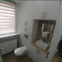 Nowoczesna, kompaktowa łazienka z białymi szafkami, podwieszaną toaletą i prostokątną umywalką nablatową. W lustrze odbija się pralka.