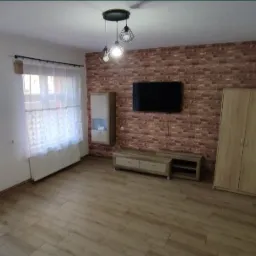 Umeblowany pokój po remoncie: ściana z cegły dekoracyjnej, drewniana podłoga, szafa, szafka RTV, telewizor, lampa wisząca z trzema kloszami, okna z białymi firankami.