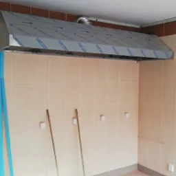 Montaż przemysłowego okapu wentylacyjnego ze stali nierdzewnej, zabezpieczonego folią ochronną, w pomieszczeniu z wykafelkowanymi ścianami i podłogą, z widocznymi szczotkami do sprzątania.