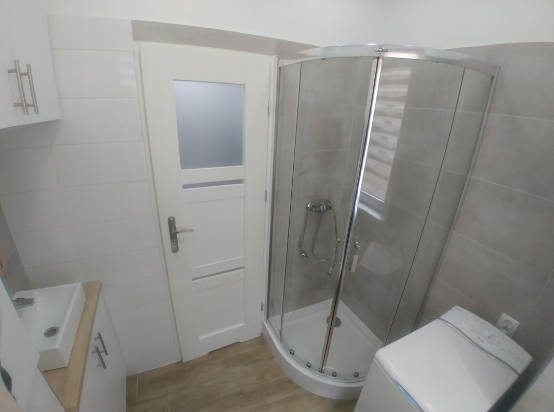 Nowoczesna łazienka po remoncie: kabina prysznicowa narożna z chromowanymi elementami, biała umywalka z szafką, białe płytki na ścianach i podłodze imitujące drewno.