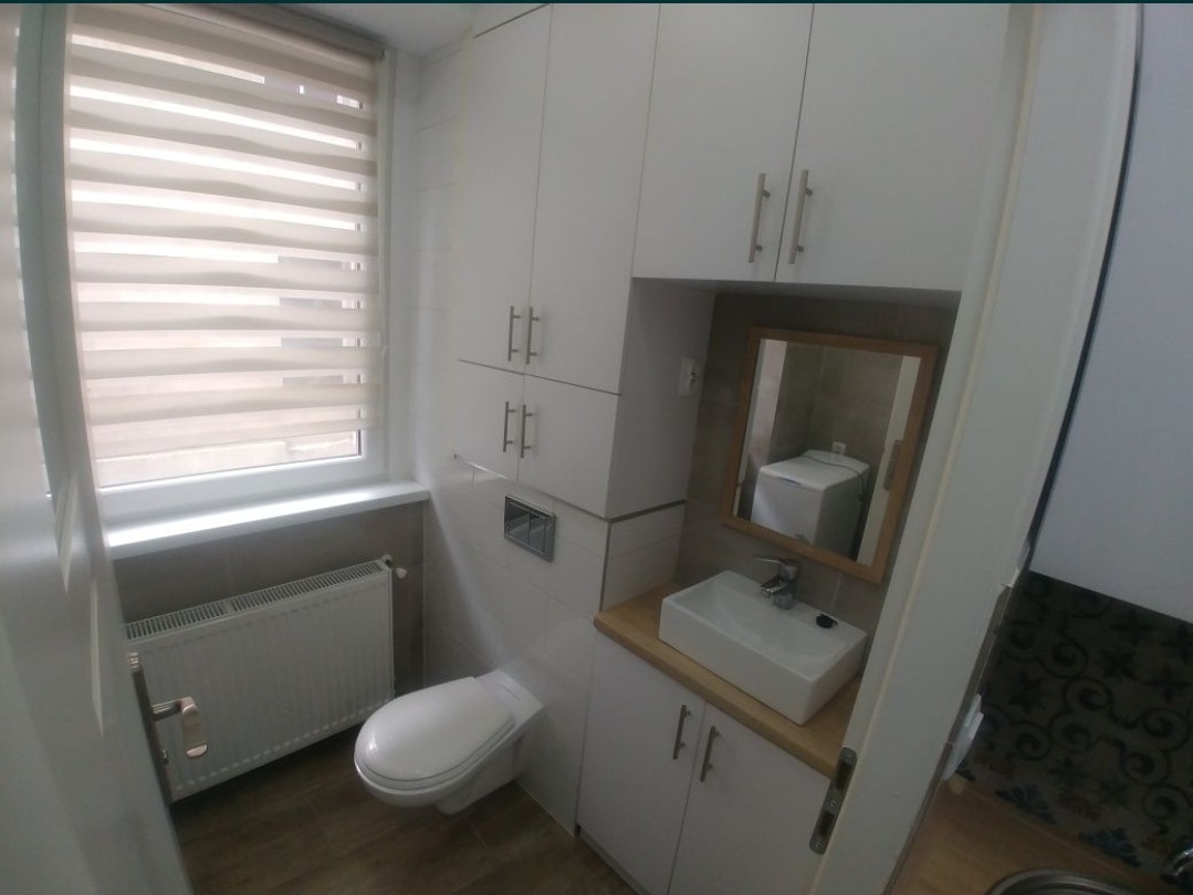 Nowoczesna, kompaktowa łazienka z białymi szafkami, podwieszaną toaletą i prostokątną umywalką nablatową. W lustrze odbija się pralka.