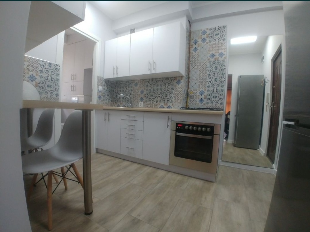 Jasna kuchnia po renowacji z białymi szafkami, płytkami w stylu patchwork nad blatem i stalowym piekarnikiem. Widoczne krzesła przy blacie i odbicie lodówki w lustrze.