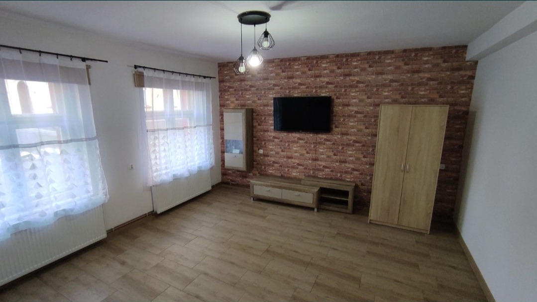 Umeblowany pokój po remoncie: ściana z cegły dekoracyjnej, drewniana podłoga, szafa, szafka RTV, telewizor, lampa wisząca z trzema kloszami, okna z białymi firankami.