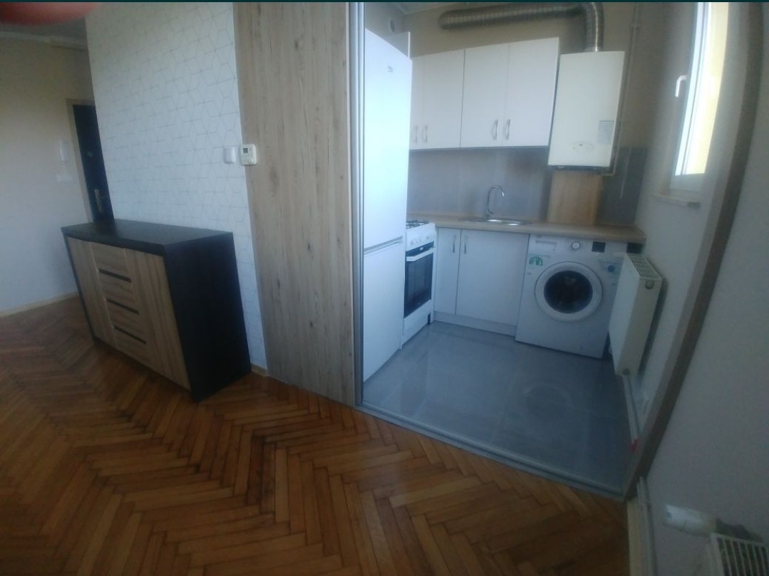 Mała kuchnia z białymi szafkami, pralką i piecem gazowym, widoczna z perspektywy pokoju z parkietem w jodełkę i komodą.