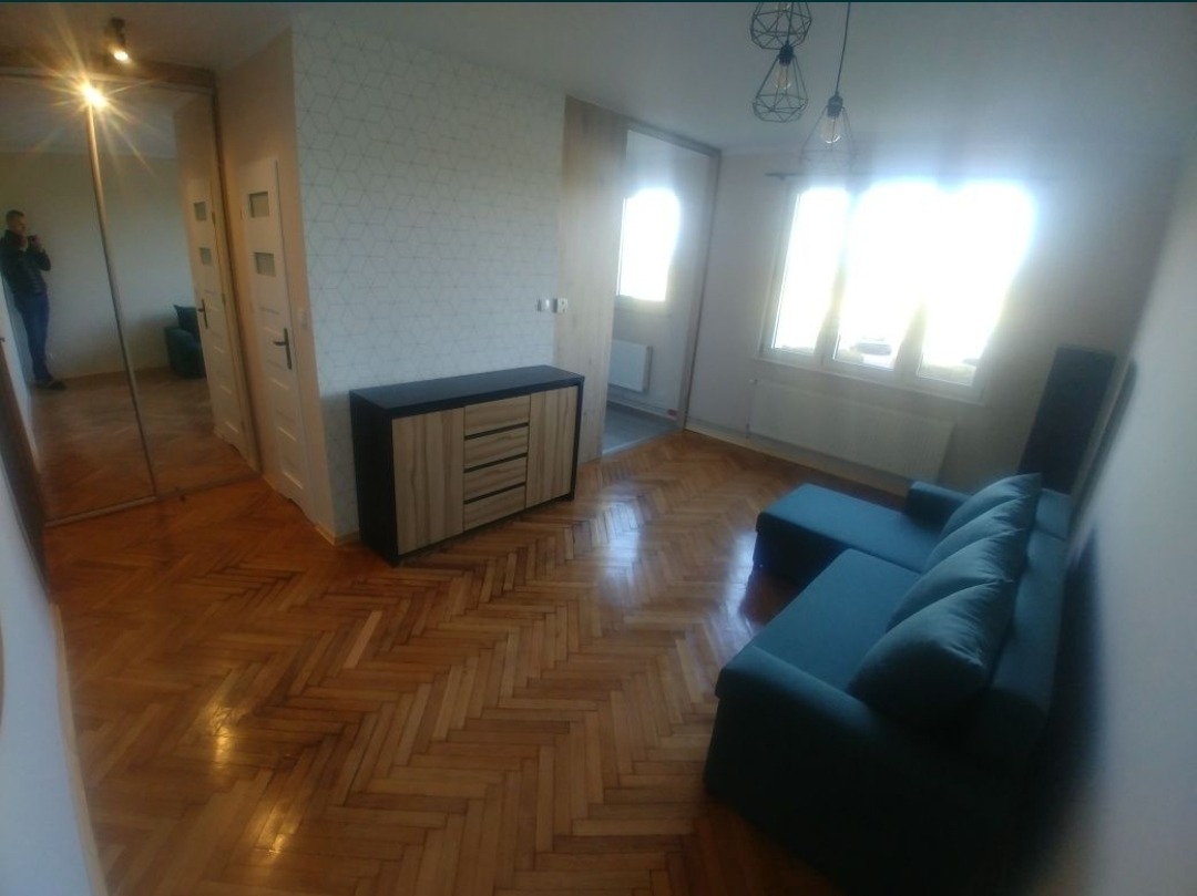 Jasne wnętrze pokoju po odświeżeniu, z parkietem w jodełkę, turkusową sofą i komodą z drewnianymi frontami.