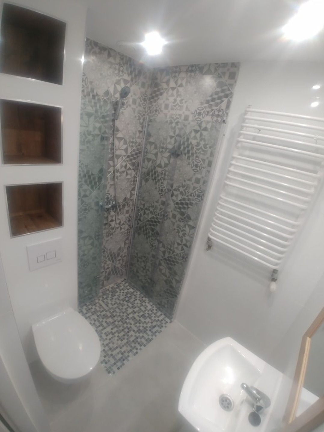 Nowoczesna łazienka z kabiną prysznicową wyłożoną mozaiką w odcieniach szarości, biała umywalka narożna, toaleta oraz grzejnik drabinkowy na ścianie.