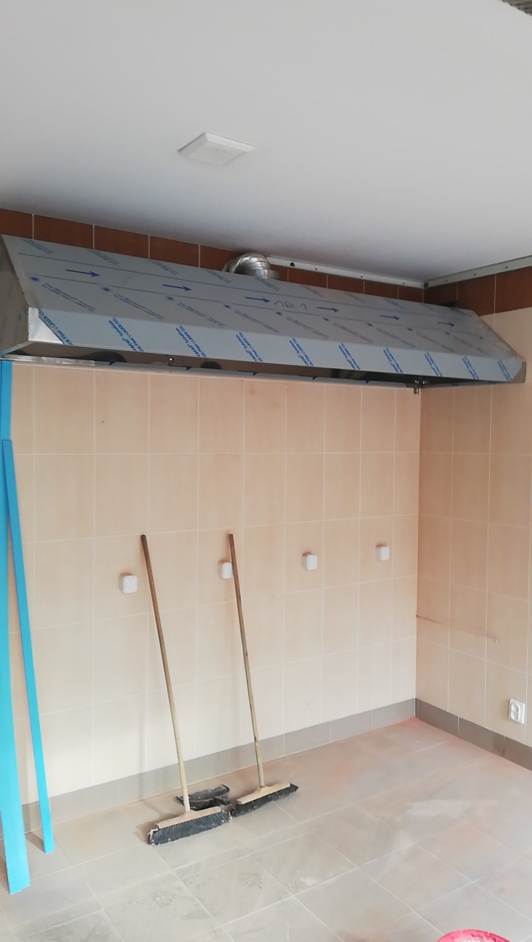 Montaż przemysłowego okapu wentylacyjnego ze stali nierdzewnej, zabezpieczonego folią ochronną, w pomieszczeniu z wykafelkowanymi ścianami i podłogą, z widocznymi szczotkami do sprzątania.