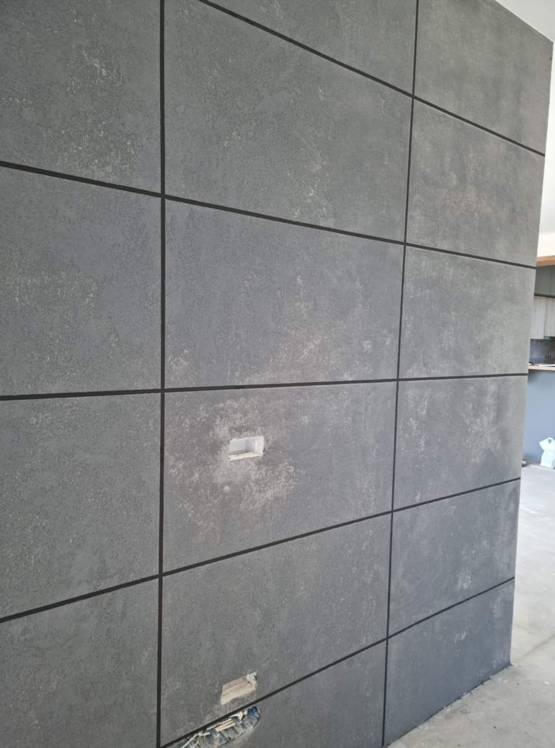 Szara ściana z betonowych płyt z fugami, widoczne otwory na gniazdka elektryczne w trakcie instalacji.