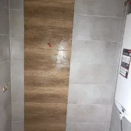 Ściana łazienki wyłożona płytkami imitującymi drewno i szarymi płytkami, widoczne przyłącza wodne i bojler.