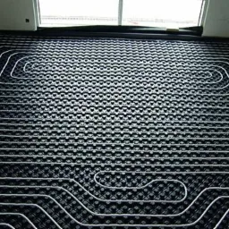 Białe rurki ogrzewania podłogowego ułożone na czarnej, strukturalnej macie w trakcie instalacji w pomieszczeniu z oknem.