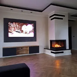 Salon z kominkiem i telewizorem w czarnej ramie, drewniana podłoga w jodełkę, czarne akcenty na suficie i kominku.