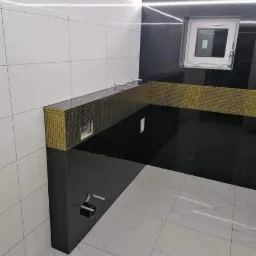 Wykończona łazienka z czarno-białymi płytkami, mozaiką w odcieniach złota nad zabudową stelaża WC i oświetleniem LED podsufitowym.