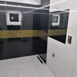 Nowoczesna łazienka z czarno-białymi płytkami, złotym pasem dekoracyjnym, podświetleniem LED i odpływem liniowym w podłodze.